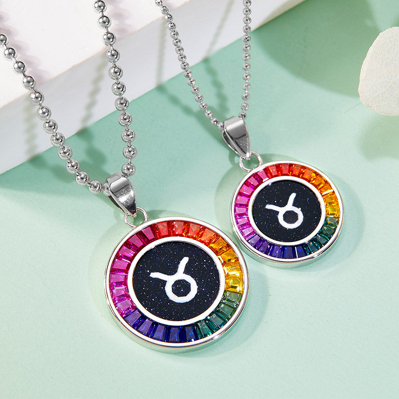 Two rainbow-colored Taurus zodiac pendants on silver chains against a light green background.